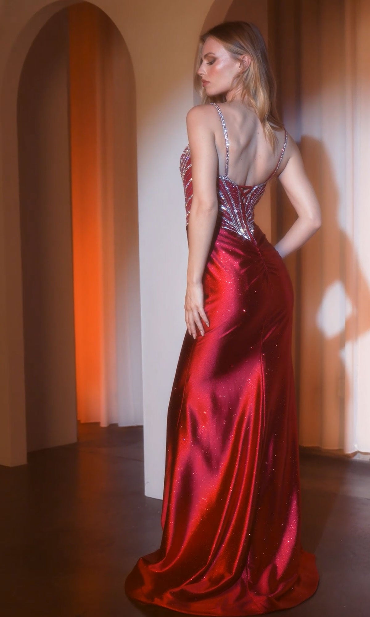 A woman in the Ladivine CC8868 Beaded-Corset Long Prom Dress, a floor-length red gown with thin straps and shimmering crystals, stands with her back to the camera in a softly lit room.